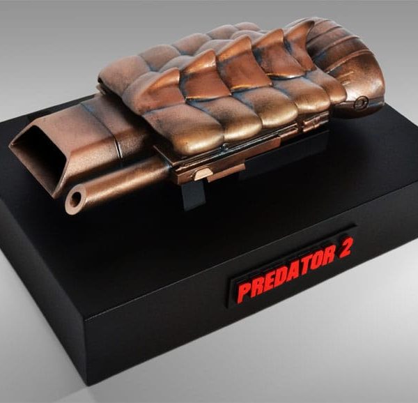 Bronze-colored Predator 2 Spear Gun prop replica on a black display base with a red Predator 2 logo